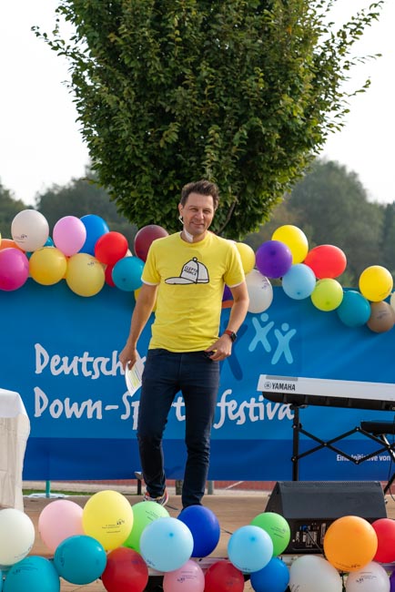 Foto: Moderator André Gatzke steht vor einem mit Bunten Ballons geschmücktem Banner.