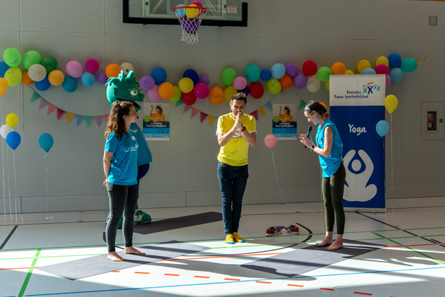 Foto: Yoga-Trainerinnen, Moderator André Gatzke und Maskottchen führen eine Übung in der Halle vor.