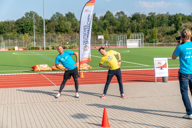 Foto: Moderator André Gatzke und ein Trainer machen Dehnübungen auf dem Sportplatz.